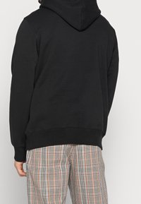 Felpa nera con tasca a marsupio e cappuccio regolabile con cordino, abbinata a pantaloni a scacchi in grigio, bianco, arancione e nero.