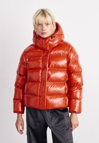 Giacca a vento rossa con finitura lucida, colletto alto e design a quadretti orizzontali. Presenta un cappuccio e una chiusura con zip frontale.