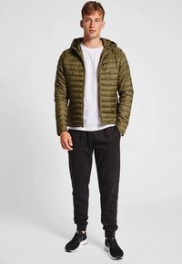 Grøn pufferjakke med hætte, lynlås og ribkanter, kombineret med en hvid skjorte og sorte bukser. Har et quiltet design.