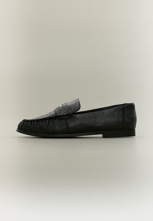 Mocassin en cuir noir avec un petit talon, un bout arrondi et des surpiqûres style mocassin à l’avant et sur les côtés.