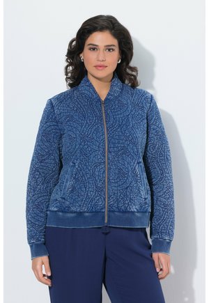 Vrouw met donker krullend haar draagt een blauw patroon vest met rits en marineblauwe broek, staand tegen een effen lichte achtergrond.