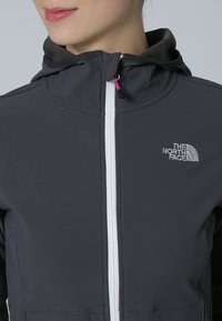 Graue Kapuzenjacke mit einem Reißverschluss, ausgestattet mit einem weißen Reißverschluss, gesticktem Logo und glatter Stoffoberfläche. Entwickelt für den Einsatz im Freien.