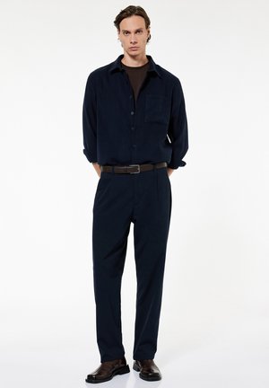 Hombre de pie con las manos detrás de la espalda, vistiendo camisa negra abotonada, pantalones negros, cinturón marrón y zapatos marrón oscuro sobre fondo blanco.