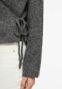 Pilkas vilnonis viršus su tekstūruotu paviršiumi, ilgomis rankovėmis ir raišteliu juosmens srityje, derinamas su baltais džinsais.