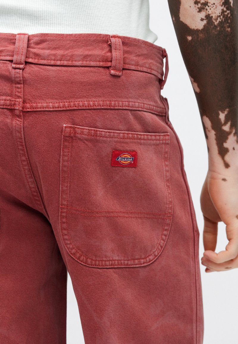Red denim pants feature a high waist, standard pockets, and a textured finish. A small red and yellow Dickies logo patch is visible.