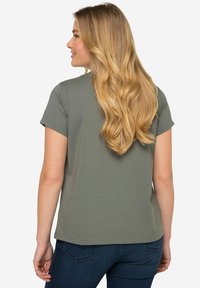 T-shirt à manches courtes, vert olive, en coton doux. Présente une coupe décontractée avec un ourlet droit. Porté avec un jean bleu foncé.