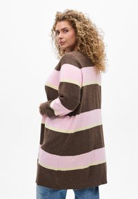Cardigan long rayé marron, rose et jaune avec une texture douce. Présente une coupe décontractée et des poignets côtelés, porté sur un jean.