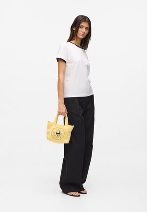 Jeune femme portant un t-shirt blanc et un pantalon noir, tenant un petit sac fourre-tout jaune avec un dessin de chat, debout sur un fond blanc.