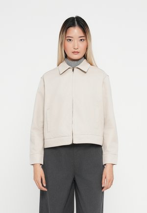 Lys beige kort jakke med spiss krage, frontglidelås og sidelommer. Glatt stoff med en avslappet passform. Brukt over en grå turtleneck.
