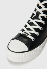 Black high-top sneaker made of synthetic leather, featuring white laces, a rounded white rubber toe cap, and a textured rubber sole with ridges.