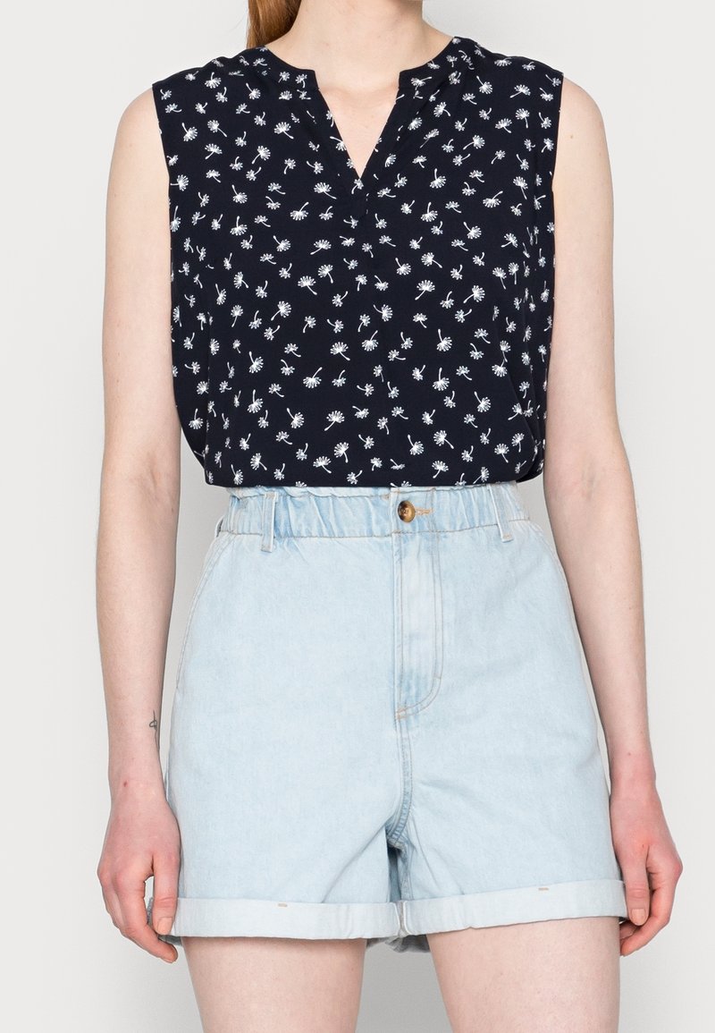 Blouse noire sans manches avec motif pissenlit blanc associée à un short en jean bleu clair taille haute avec ourlets retournés.