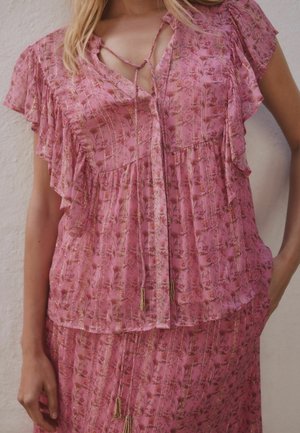 Frau trägt eine rosa Bluse mit floralem Muster, kurzen Rüschenärmeln und Quastenbändern am Ausschnitt, kombiniert mit einem passenden Rock.