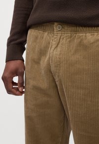 Pantalon en velours côtelé de couleur beige chaud, avec des côtes verticales, une fermeture à bouton et une taille élastique. Détails de tissu texturé visibles.