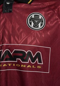 Bordo värvi spordisärk musta ja kuldse kraega, must-valge ümmargune meeskonna sümbol "XXIII" ja paks must sponsoritekst "WARM NATIONALS."