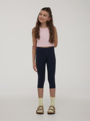 Giovane ragazza in piedi, sorridente, indossa un top rosa senza maniche, leggings capri blu navy, calzini gialli e sandali marroni su uno sfondo uniforme.