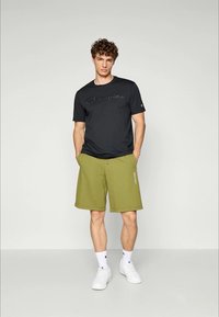 Junger Mann steht mit den Händen in den Taschen, trägt ein schwarzes Champion-T-Shirt, olivgrüne Shorts, weiße Socken und weiße Sneakers vor einem einfarbigen Hintergrund.