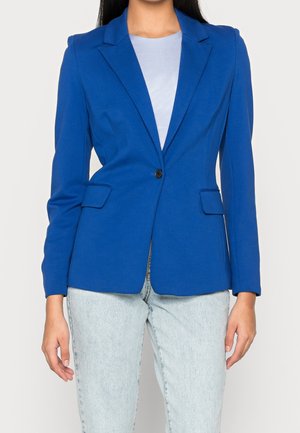 Femme portant un blazer bleu ajusté avec un bouton noir, sur un haut bleu clair et un jean délavé clair, debout devant un fond uni.