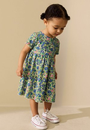 Unge barn med fletninger iført en blomsterkjole og hvide sneakers, kigger nedad mod en ensfarvet beige baggrund.
