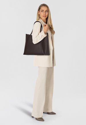 Sac fourre-tout en cuir texturé marron foncé avec un design épuré et angulaire. Doté de deux poignées et d'un intérieur spacieux. Porté avec un costume de couleur claire.