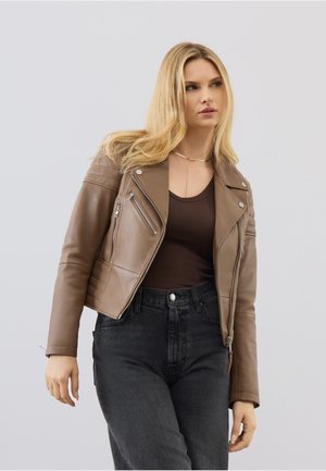 Blond kvinde iført en brun læderjakke, mørkebrun top og sorte højtaljede jeans, stående foran en ensartet lys baggrund.