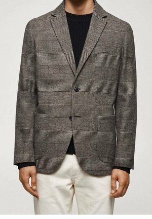 Hombre vestido con un blazer a cuadros marrón y negro sobre un suéter negro y pantalones blancos, de pie con los brazos relajados a los lados.