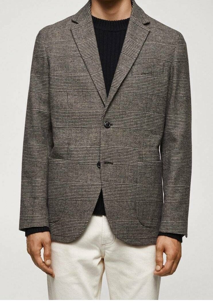 Homme portant un blazer à carreaux marron et noir sur un pull noir et un pantalon blanc, debout, les bras détendus le long du corps.