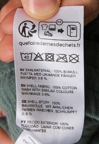 Étiquette avec des instructions d'entretien imprimées en plusieurs langues : 100 % coton, inclut des symboles de lavage et des informations sur le rétrécissement.