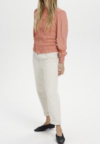 Persikofärgad blus med smockning och långa ärmar, vita uppvikta byxor och svarta platta skor. Slätt tyg med en subtil textur.