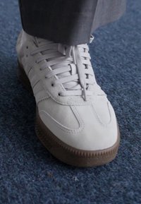 Fehér sneaker, amely velúrból és hálós anyagból készült, texturált barna gumitalppal, fűzős kialakítással és diszkrét varrási részletekkel rendelkezik.