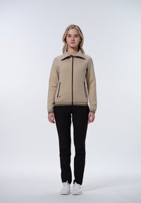 Beige zipová bunda s límcem, černými akcenty a dekorativním vzorem na manžetách. Na sobě má černé kalhoty a bílé tenisky.