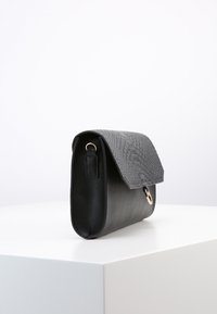 Černá kožená kabelka s texturovaným klopovým víkem a zlatým uzávěrem, umístěná na bílé podstavě na jednoduchém bílém pozadí.