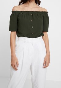 Olivi vihreä olkaimeton pusero, jossa on lyhyet hihat, rypistetty kaula-aukko ja kolmesta napista koostuva etulukko. Yhdistetty valkoisiin korkeavyötäröisiin housuihin.