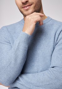 Maglione azzurro chiaro a coste con scollo rotondo e maniche lunghe, realizzato in un tessuto testurizzato. Il modello sta poggiando il mento sulla mano.