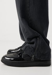 Black patent leather sneakers with a textured finish, featuring lace-up front and a thick black sole. Pants are dark, with a zippered ankle detail.