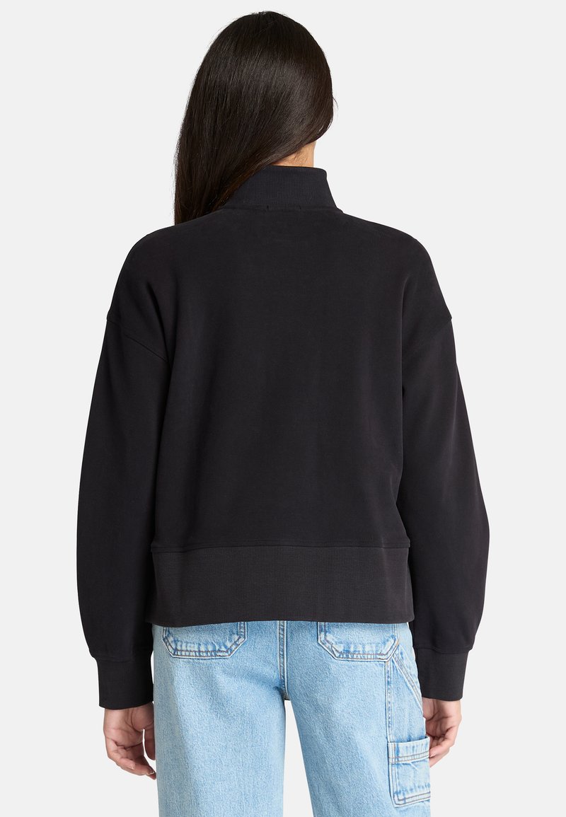 Felpa nera cropped con colletto alto, spalle basse e polsini a costine. Presenta una texture liscia e una vestibilità rilassata, abbinata a jeans chiari.