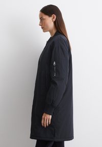 Veste bomber noire avec une coupe longue et décontractée, manches à bords côtelés, poche latérale zippée et texture lisse. Présentée avec une fermeture éclair sur le devant.