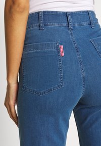 Blå denimjeans med hög midja, med en bakficka och en röd och vit rutmönstrad etikett. Mjuk textur och klassisk design.