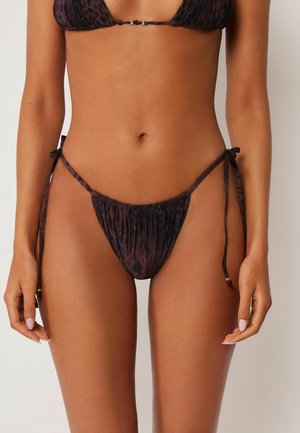 Femme portant un bikini à imprimé léopard violet foncé avec un bas à nouer sur les côtés, debout devant un fond clair uni.