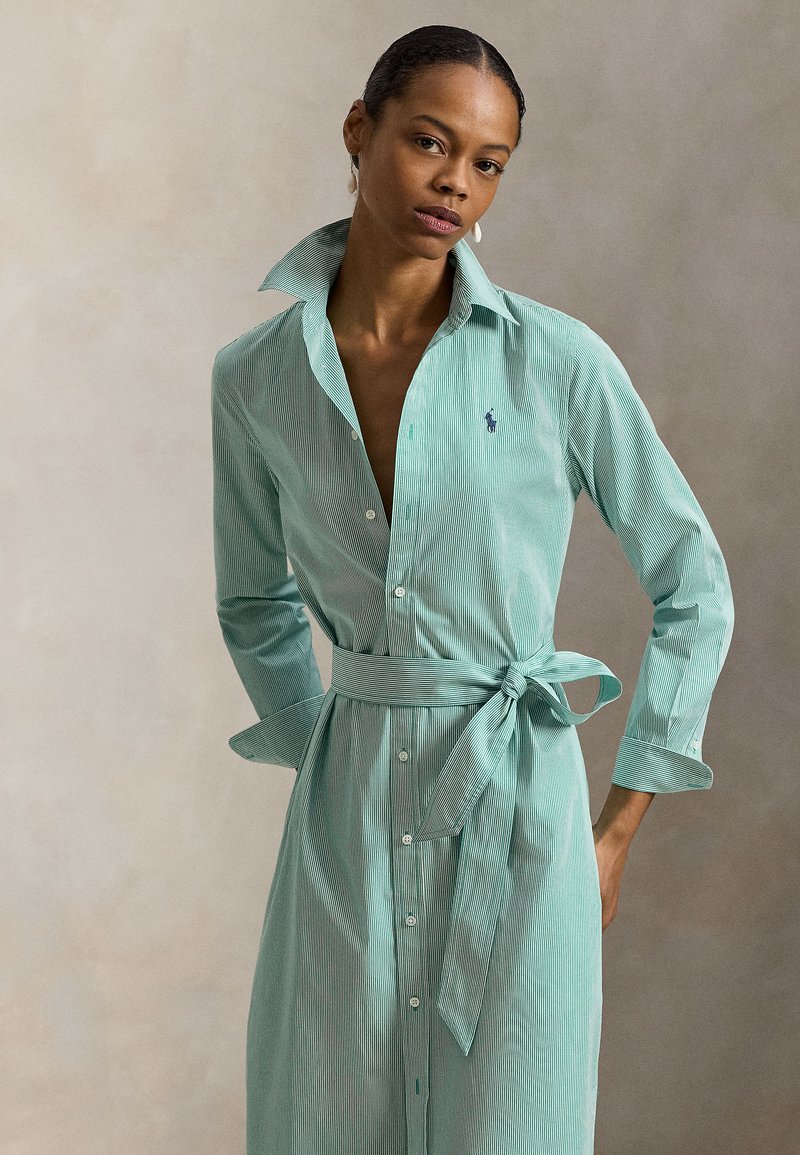 Φόρεμα πουκάμισο με πράσινες και λ ευκές ρίγες, εφαρμοστή γραμμή, κλείσιμο με κουμπιά, μακριά μανίκια και δεμένη μέση. Το γιακάς είναι όρθιος και το ύφασμα είναι βαμβακερό.