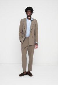 Beige double-breasted suit jacket with wide lapels, paired with a light blue shirt and beige trousers. Dark brown slip-on shoes.