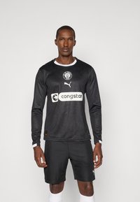 Czarna koszulka piłkarska z długim rękawem i białymi akcentami, z logo FC St. Pauli, logo Pumy oraz logo sponsora na klatce piersiowej. Noszona z czarnymi szortami.