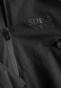 Zwarte ritssluiting jas van een gestructureerde stof, gemarkeerd met "SUB.5 //01 2" in een subtiele print. Inclusief duurzame rits hardware.