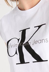 Bílý bavlněný tričko s velkým černým logem "CK" a textem "Calvin Klein Jeans" v menším šedém písmu níže. Měkká textura, kulatý výstřih.