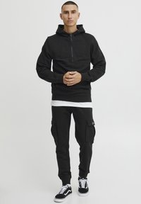 Svarte zip-up hettegensere med kengurulomme, parret med svarte cargo-bukser med sidelommer. Modellen har på seg svarte joggesko med hvite såler.