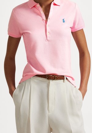 Person iført en lyserød kortærmet polo skjorte, der er stoppet ind i højtaljede cremefarvede bukser med et brunt flettet bælte.