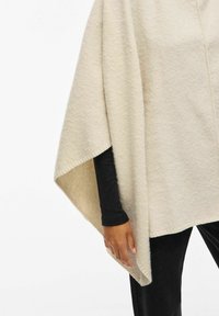 Cremefarvet uldponcho med en blød tekstur, der har en løs pasform og afrundede kanter. Beklædningen falder over en sort langærmet top.