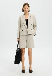 Blazer doppiopetto grigio chiaro con bottoni marroni, pantaloni corti abbinati e una borsa a rete nera. Top nero e scarpe slip-on in rete.
