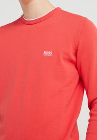 Roter Pullover aus weichem Strickstoff. Mit einem runden Ausschnitt, der einen kontrastierenden Innenbesatz und ein kleines weißes Logo auf der Brust aufweist.