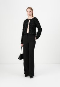 Veste cropped noire texturée avec un motif floral, devant ouvert avec fermeture à nouer, associée à un pantalon noir taille haute et un sac à main.