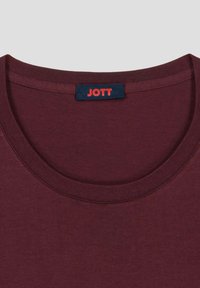 Camiseta de algodón burdeos con escote redondo y ribetes acanalados; presenta una etiqueta azul con la inscripción "JOTT" en rojo. Textura suave, diseño casual.